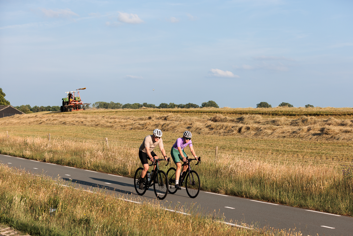 Twee wielrenners fietsen op het fietspad langs een weiland waar een boer het gras maait op zijn trekker.